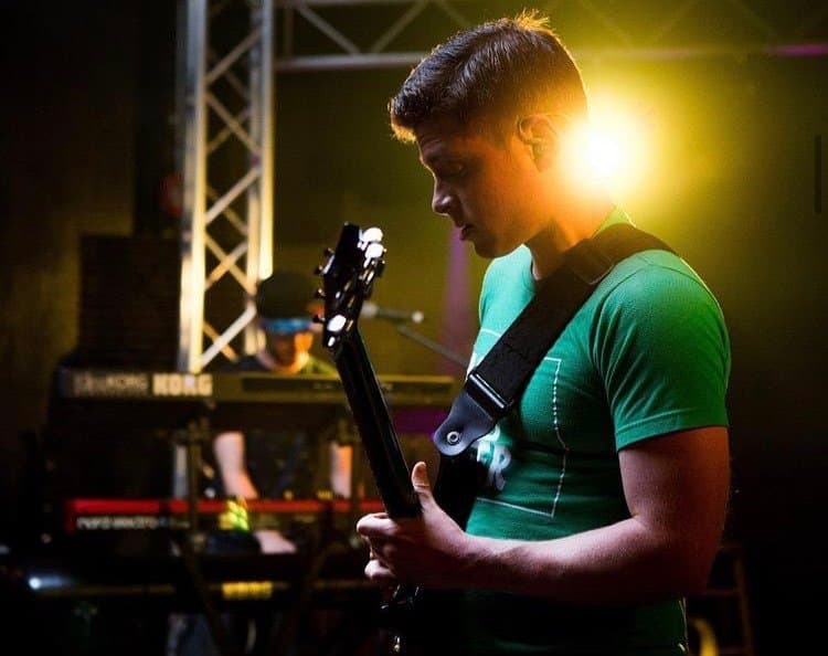 Julian Goddard playing guitar under warm stage lights with a keyboard player behind him