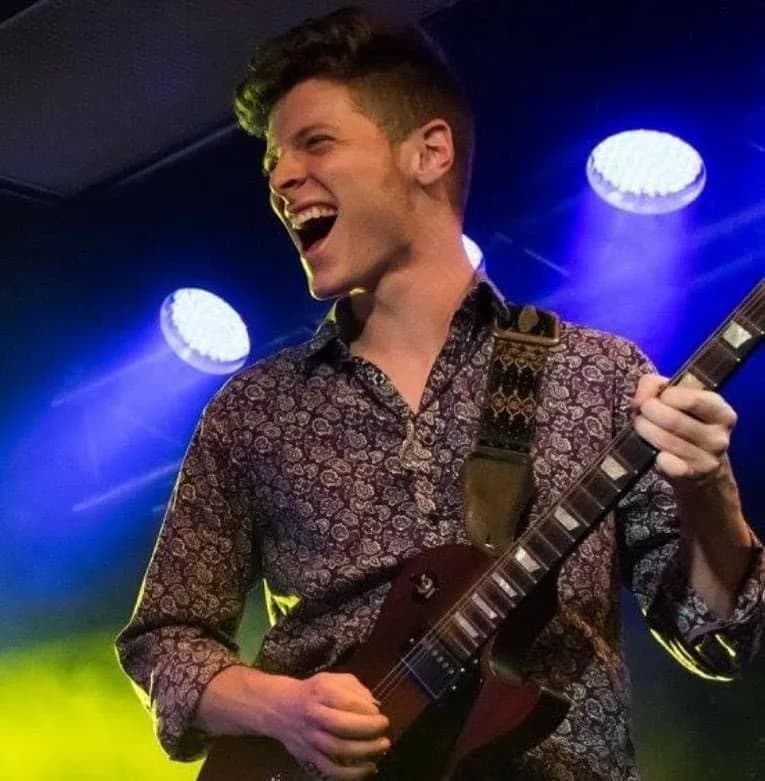 Julian Goddard grinning mid-song under blue stage lights, playing electric guitar