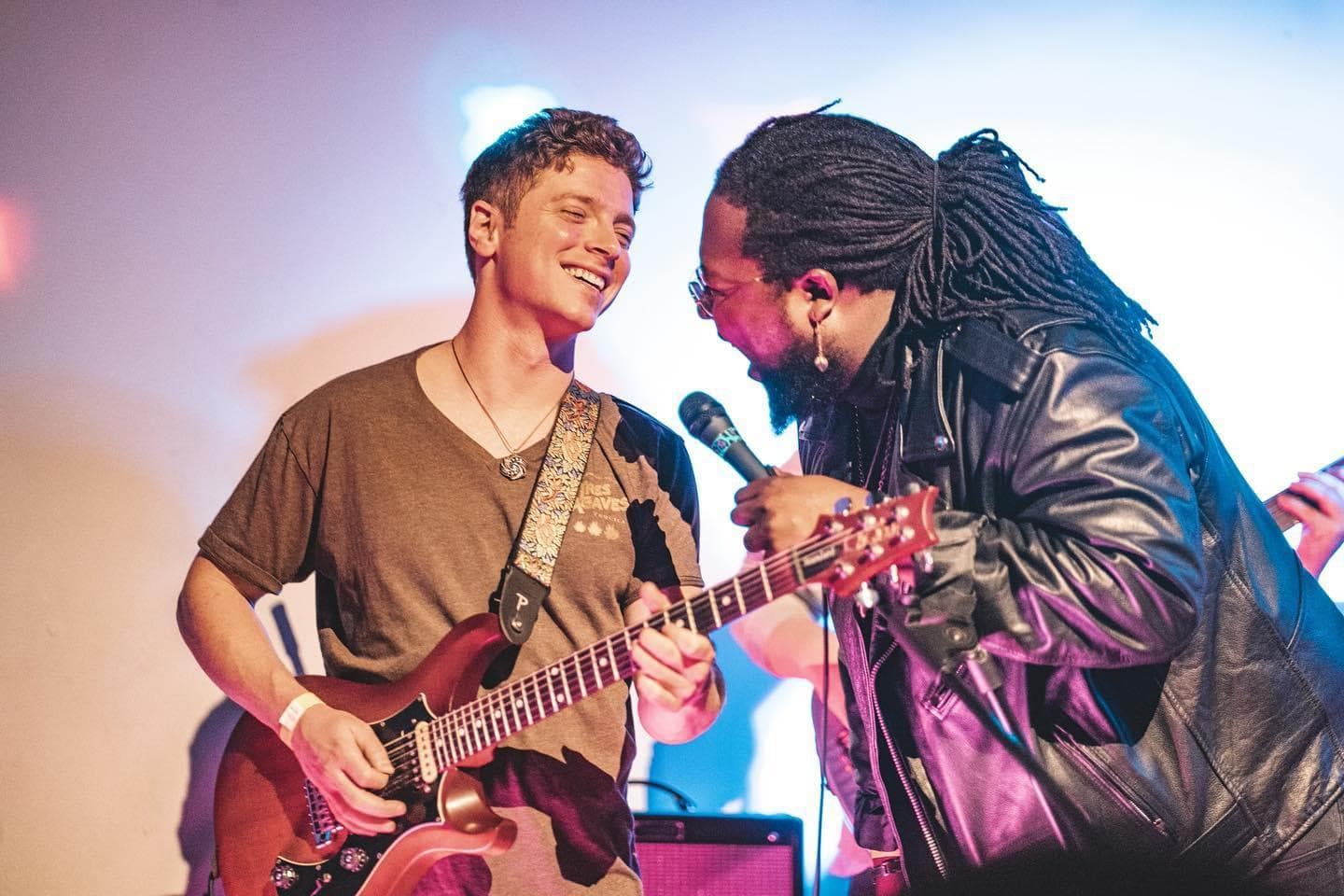 Julian Goddard sharing a microphone with a singer on stage under blue light