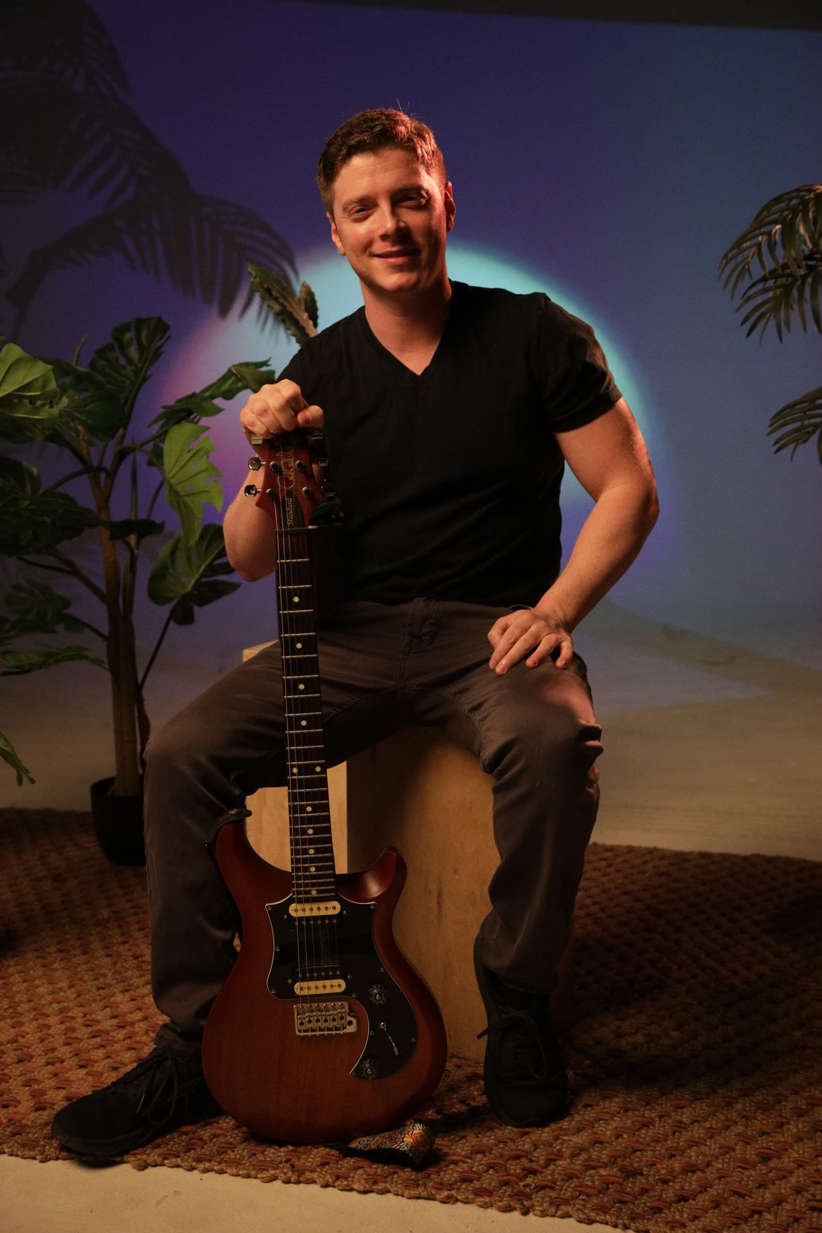 Guitar teacher Julian Goddard seated with his guitar, smiling, in a warm studio portrait