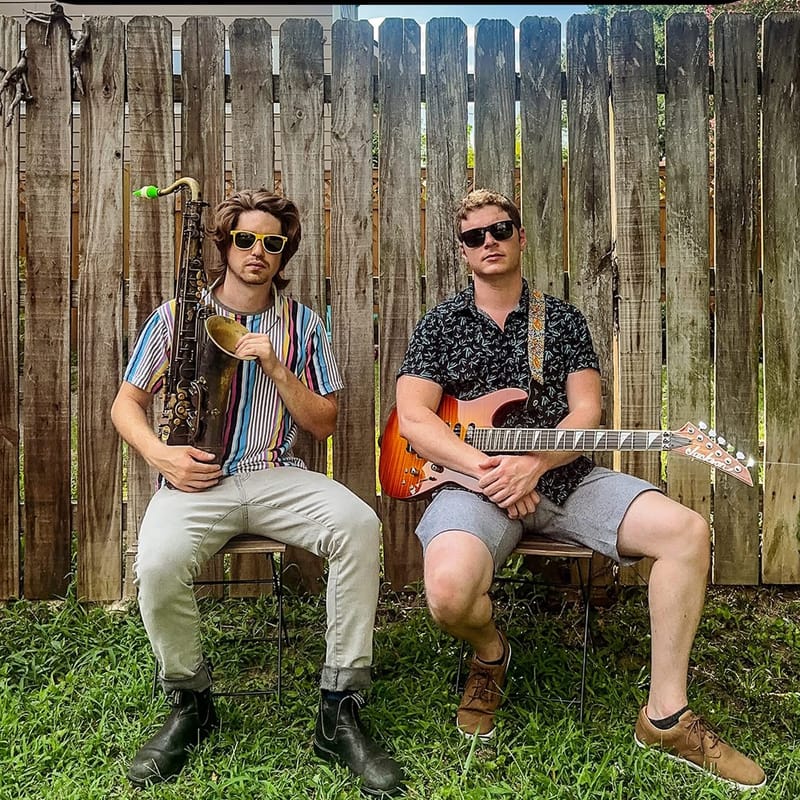 JJ Express, Julian Goddard's band: two musicians in sunglasses are seated in front of a wooden fence, one holding a saxophone and the other an electric guitar.